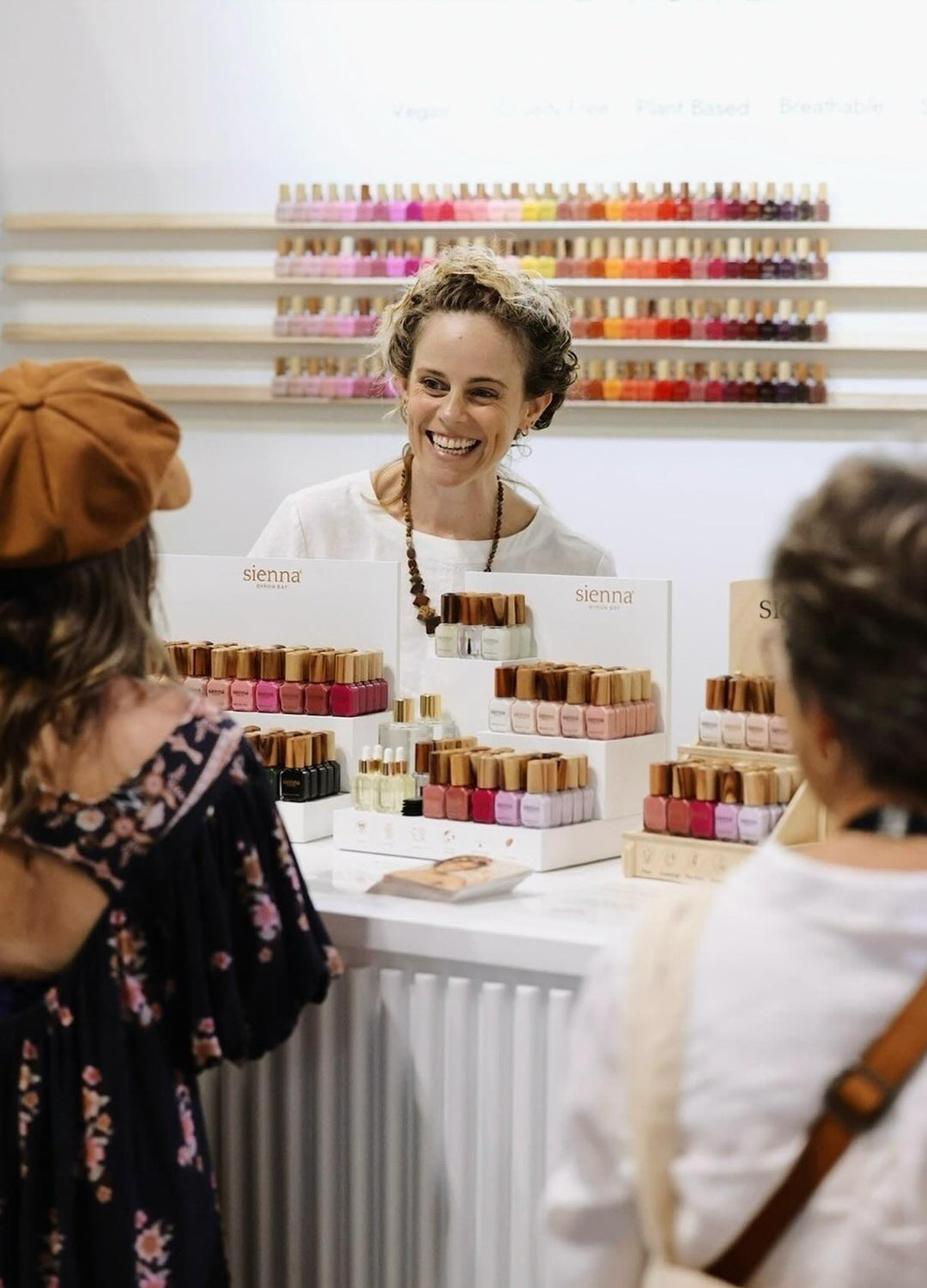 A smiling woman standing behind a retail counter filled with rows of Sienna Byron Bay nail polish.