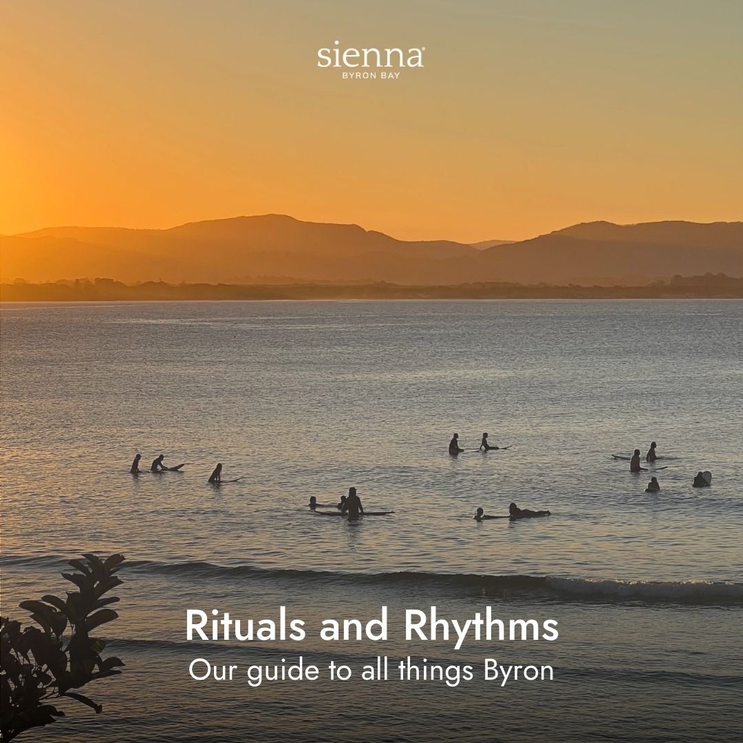 Surfers float in a calm ocean at sunset, with a mountain silhouette in the background and text reading "Rituals and Rhythms: Our guide to all things Byron.