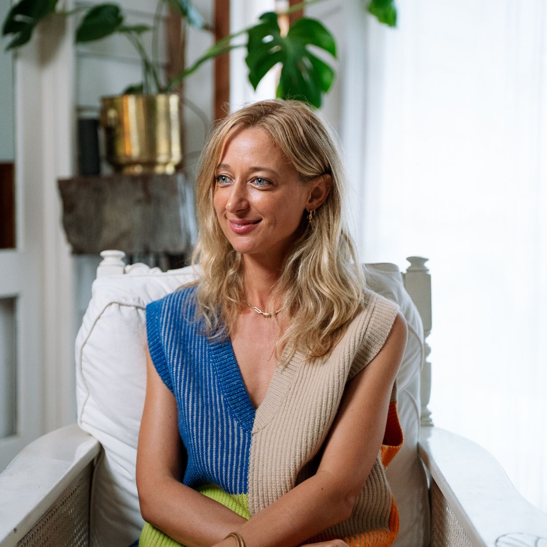 A blonde woman in a blue and beige knitted vest sitting in a white armchair next to a potted Monstera plant.