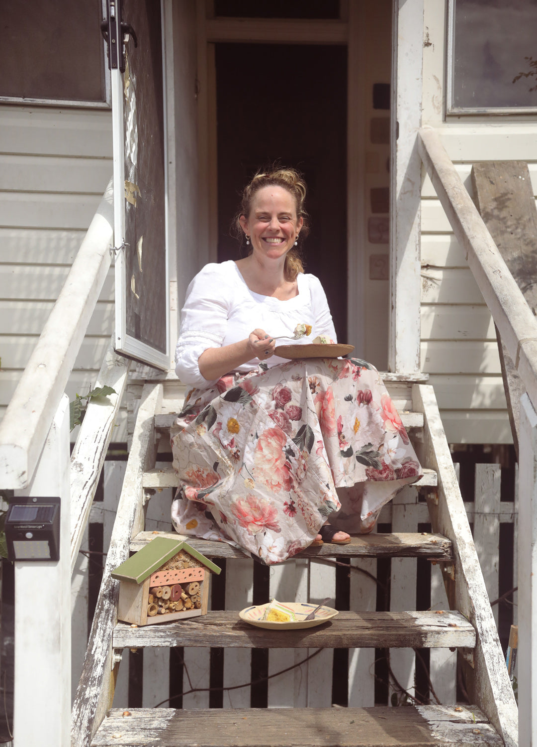 A smiling woman in a floral skirt sits on porch steps eating cake next to a bee hotel.