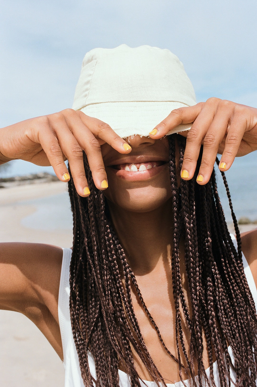 young woman wearing yellow nail polish by sienna and holding her bucket hat at the beach