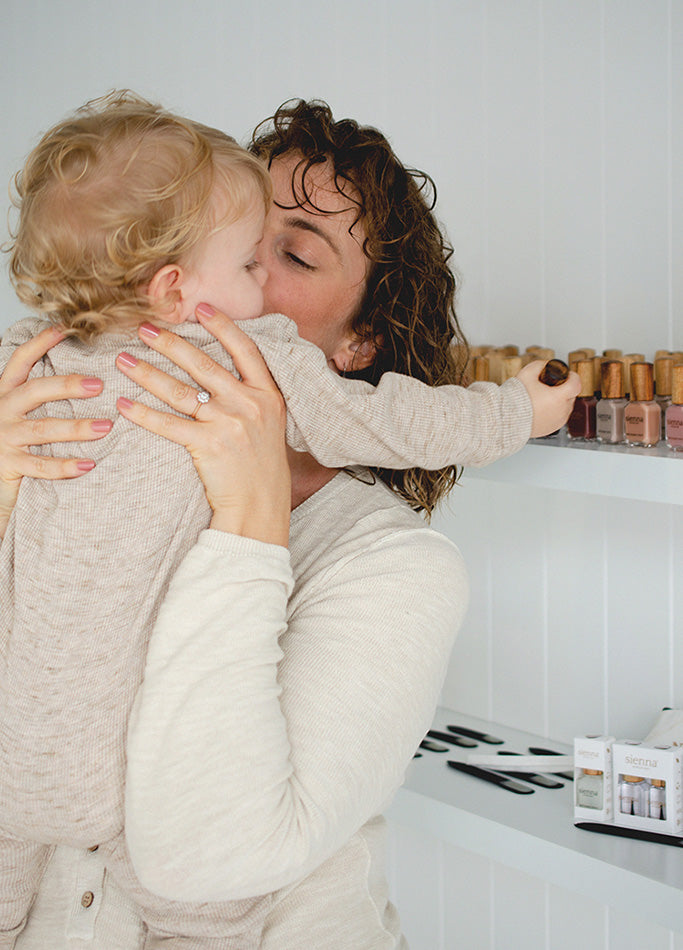 Woman with fair skin holding baby and kissing his cheek wearing Stone non-toxic nail polish by Sienna Byron Bay, and baby grabbing bottle from white shelf with bottles of non-toxic nail polish by Sienna Byron Bay.