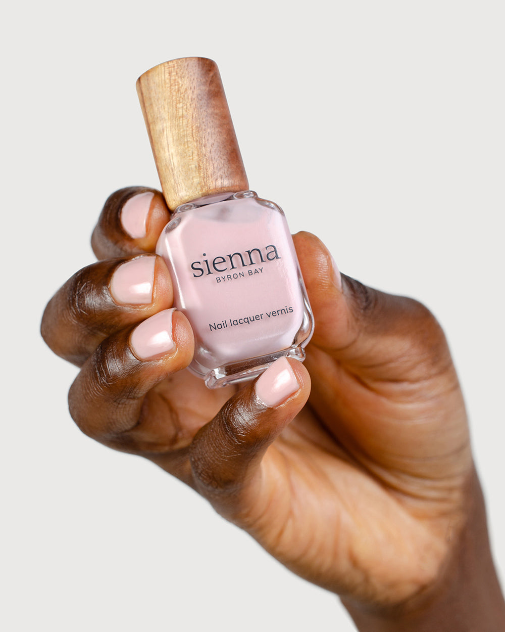 Dark skin tone hand with Amour soft pink crème nail colour and strong and healthy natural nails holding a closed bottle of Sienna Nail lacquer vernis
