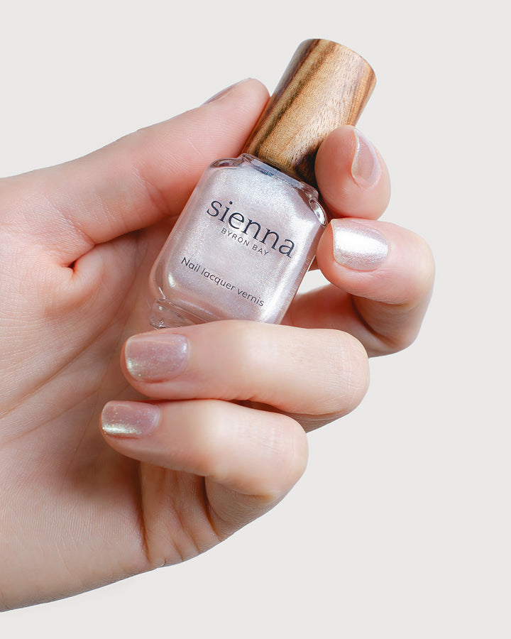 Glimmer holographic alabaster shimmer nail polish hand swatch on fair skin tone, holding a Sienna bottle