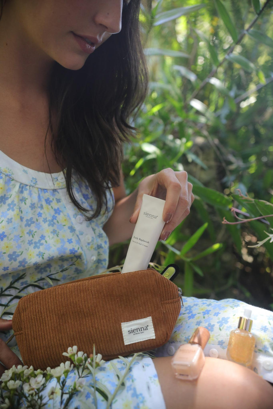 Woman holding a sienna brown corduroy bag with hand cream, nail serum and cuticle oil surrounded by greenery.