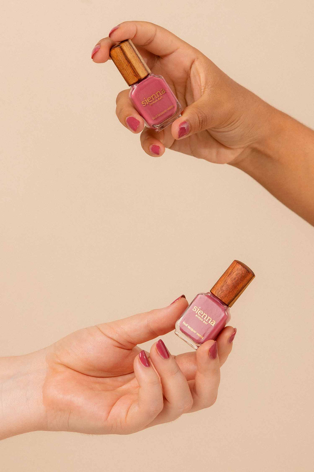 Fair skin and medium skin tone hands wearing Heartspace raspberry sorbet nail polish and holding bottles with timber lids by Sienna.