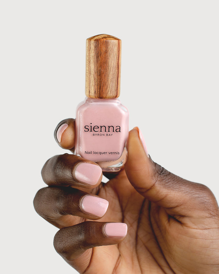 Dark skin tone hand holding a Sienna Byron Bay nail lacquer vernis bottle with signature timber cap in shimmer colour Magic. Nails are strong, and healthy and painted in shimmery Magic pink.