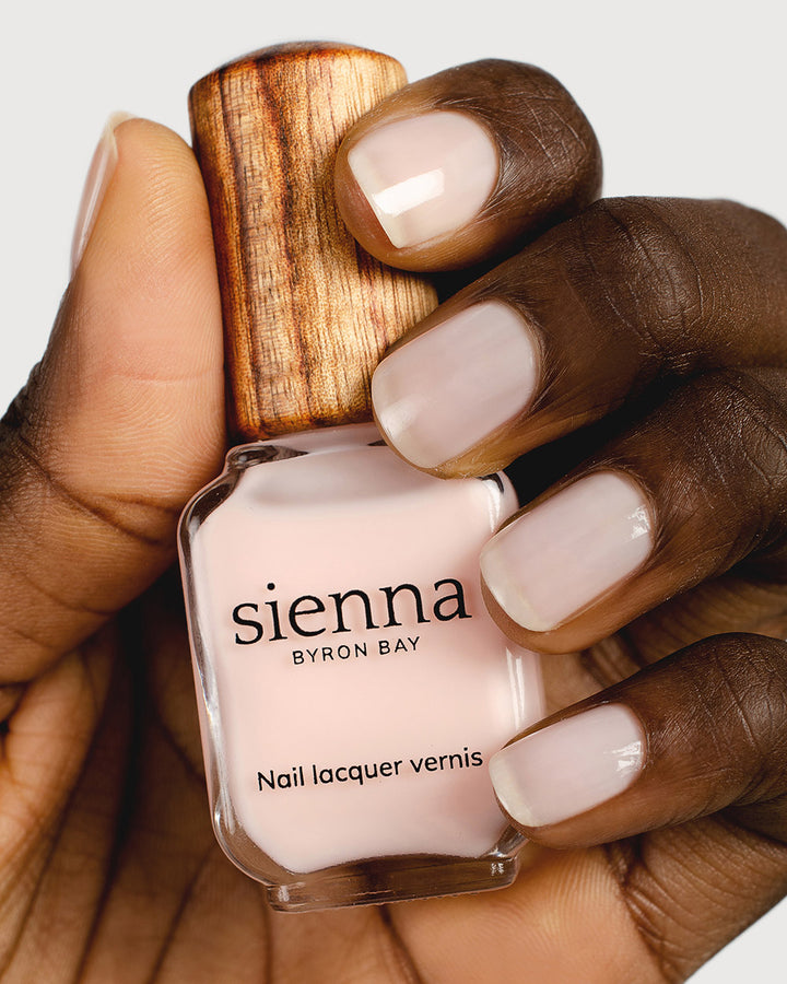 Dark skin tone hand holding a Sienna Byron Bay nail lacquer vernis bottle with signature timber cap in colour Peace. Nails are strong, and healthy and painted in sheer Peace pink.