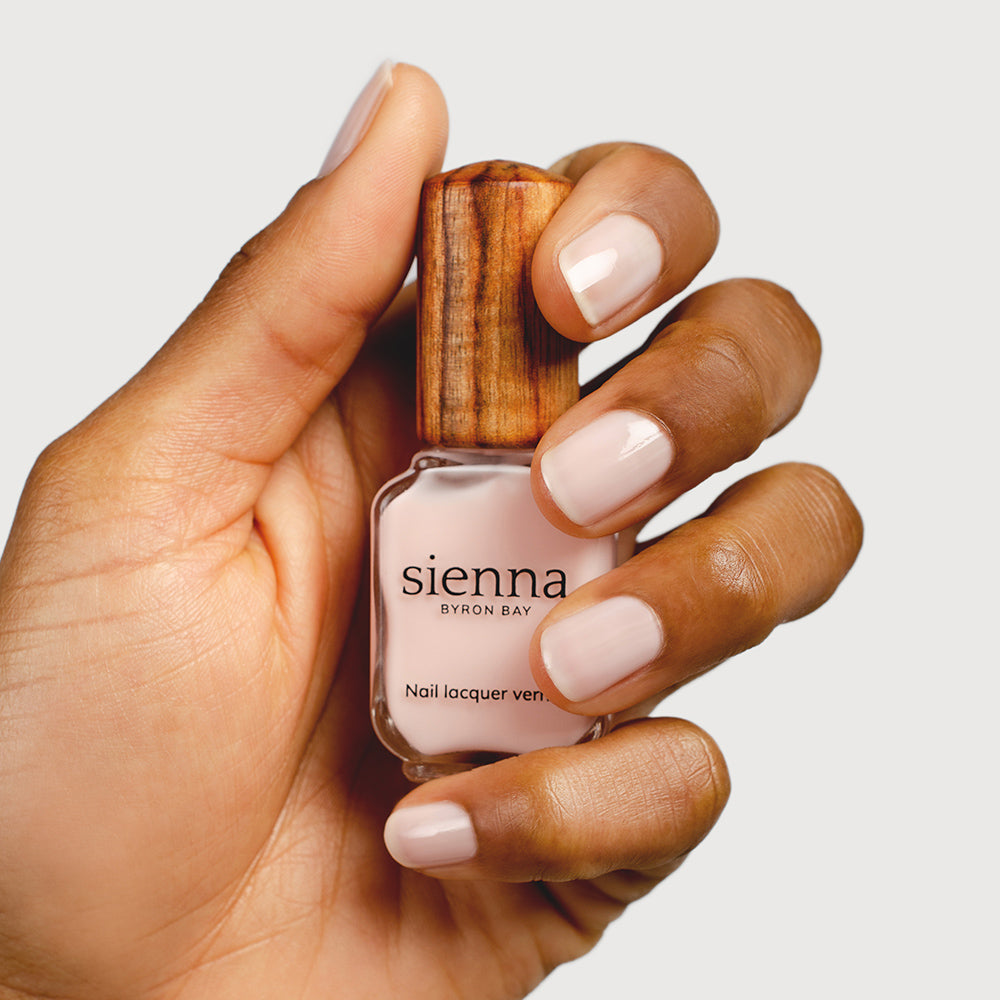Medium skin tone hand holding a Sienna Byron Bay nail lacquer vernis bottle with signature timber cap in colour Peace. Nails are strong, and healthy and painted in sheer Peace pink.