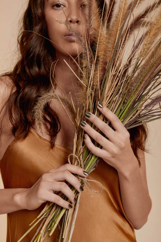 medium skin tone model wearing gold dress and black crushed crystal nail polish by sienna and holding dried flower bouquet on beige background