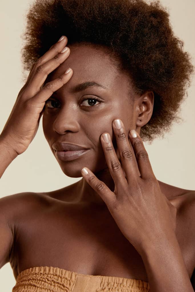 dark skin model holding her hands to her face wearing pink nude crushed crystal nail polish by sienna on a beige background