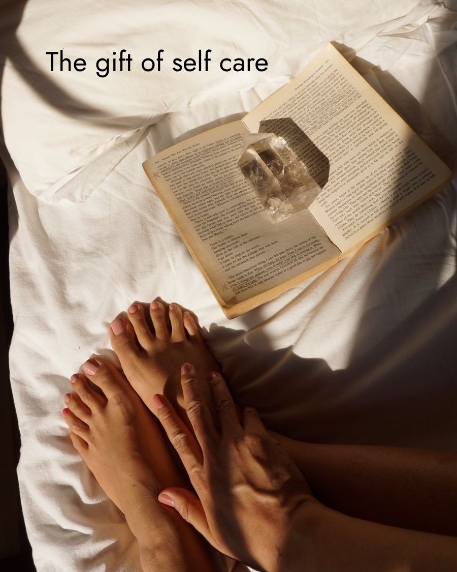 Open book with a stone on a white surface, accompanied by feet and hands, with text 'The gift of self care'.
