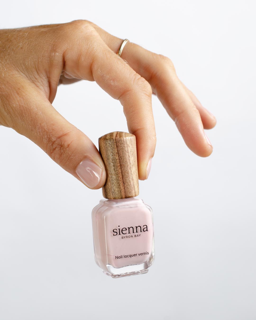 Fair skin tone hand with Amour soft pink crème nail colour and short, healthy natural nails holding the Amour bottle with signature Sienna timber cap.