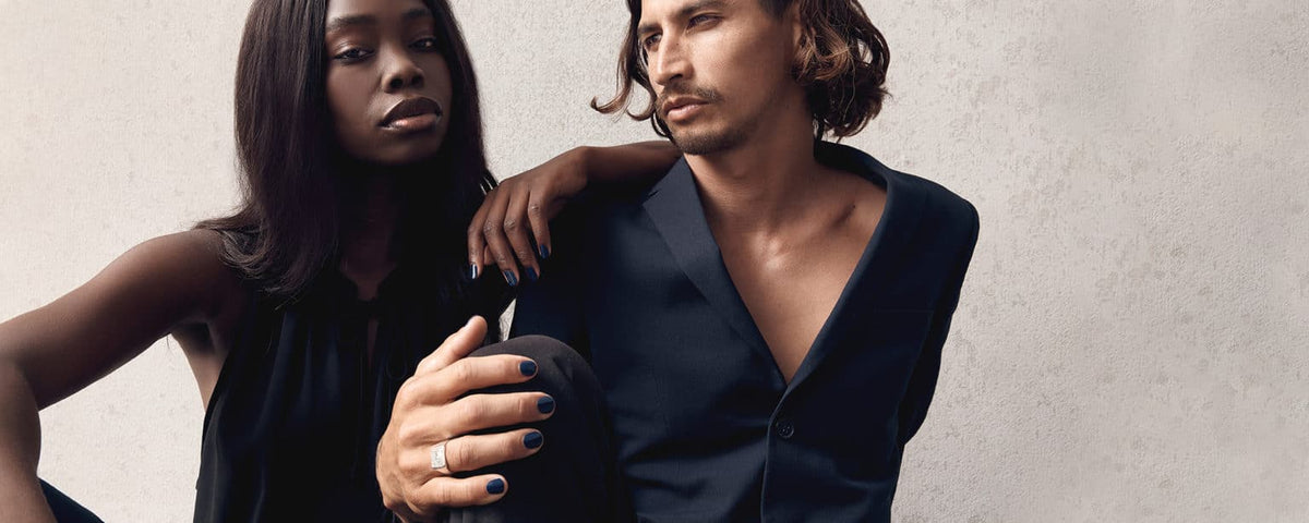 dark skin young woman and fair skin young man with long hair wearing chic black and navy shirts wearing navy blue nail polish by sienna with concrete wall on the background