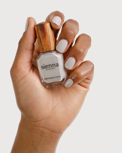 Light cool-grey nail polish hand swatch on medium skin tone holding sienna bottle