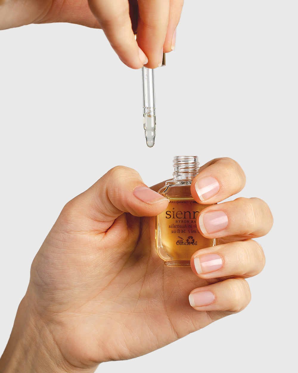 fair skin tone hand holding open bottle of cuticle oil  other hand holding dropper above the bottle