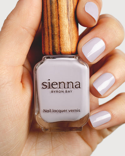 Pastel purple-grey nail polish hand swatch on fair skin tone holding a sienna bottle 