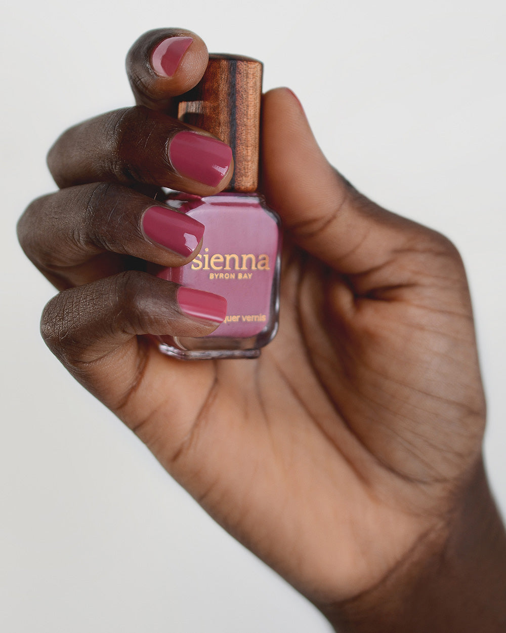 Dark skin hand wearing Heartspace raspberry sorbet nail polish and holding bottle with timber lid by Sienna Byron Bay