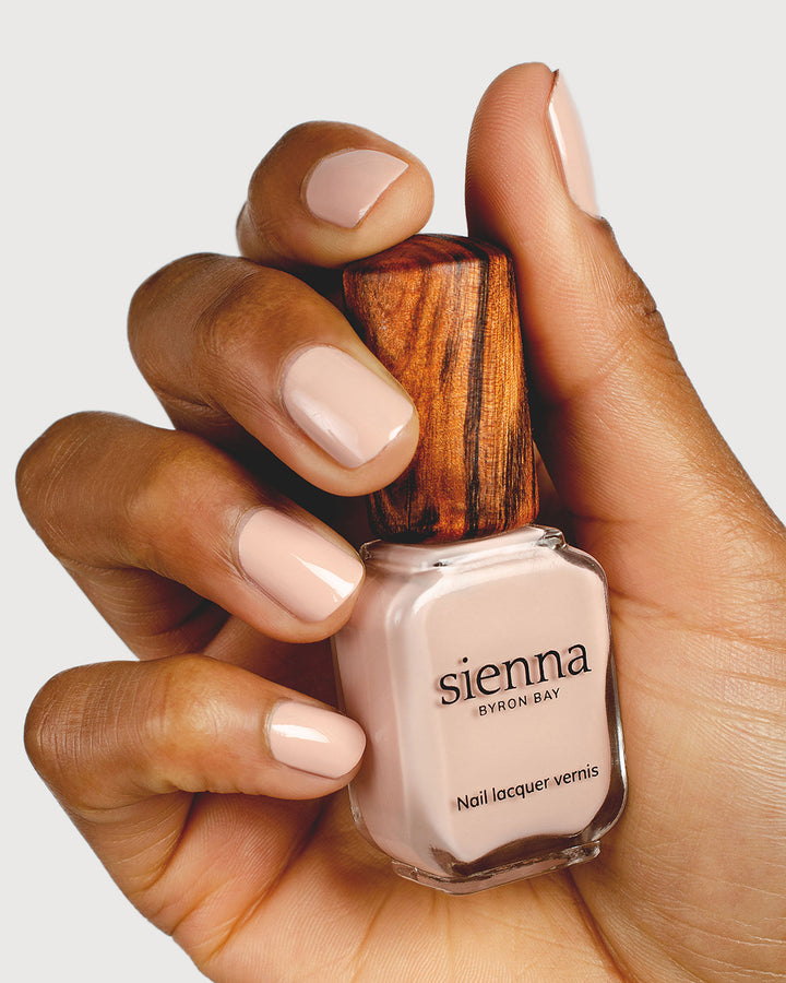 Soft neutral-pink nail polish hand swatch on medium skin tone holding sienna bottle close-up