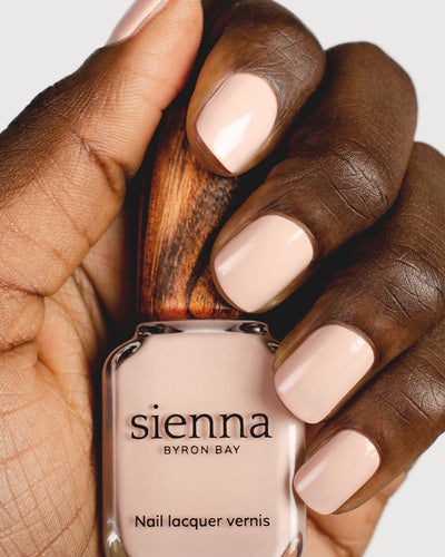 Soft neutral-pink nail polish hand swatch on dark skin tone holding sienna bottle close-up