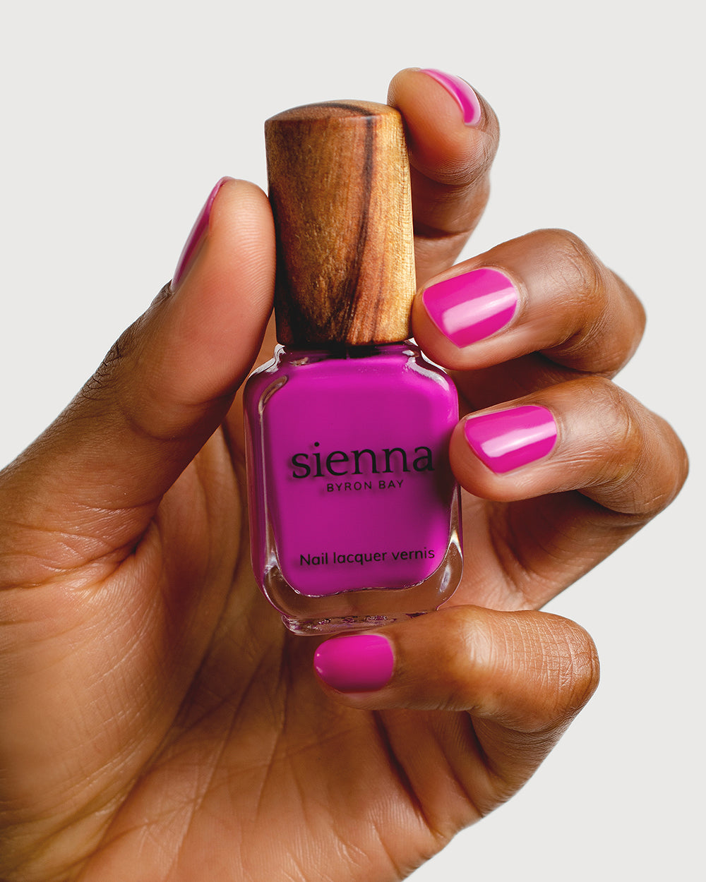 Bright magenta nail polish hand swatch on medium skin tone holding sienna bottle close-up