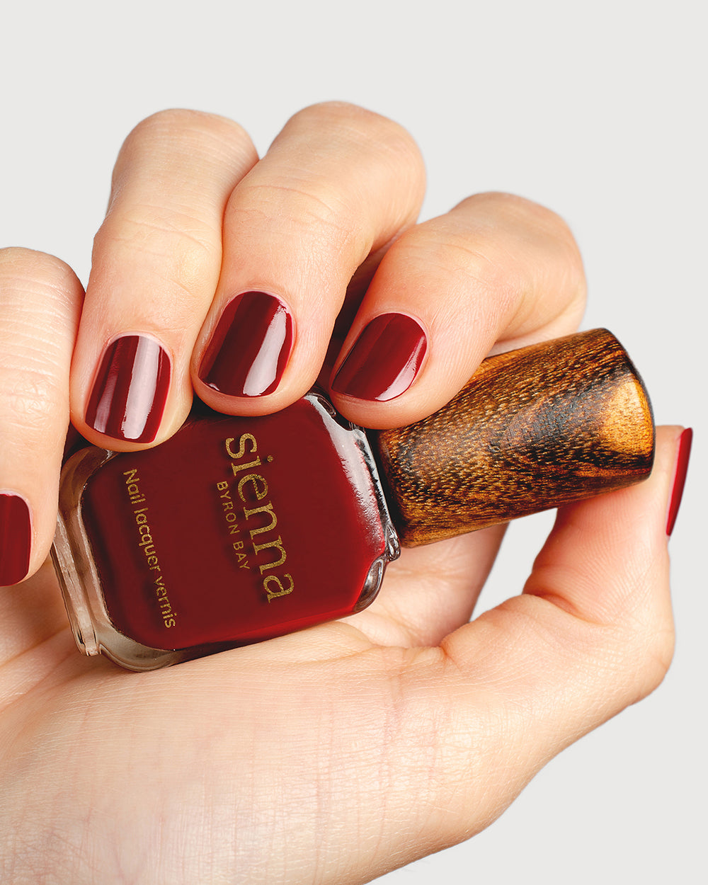 Organic mid-tone red nail polish hand swatch on fair skin tone up-close holding sienna bottle
