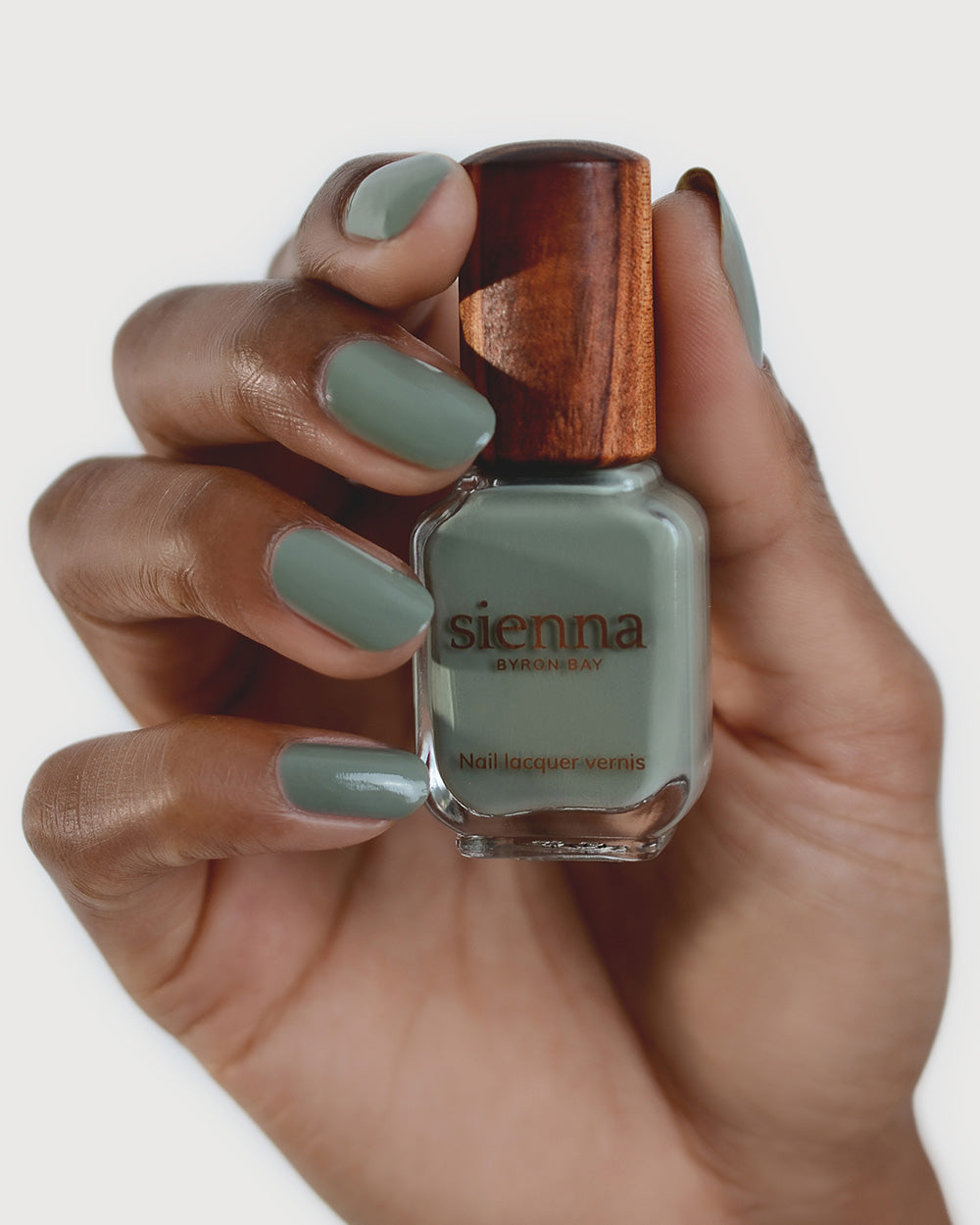 Medium tanned skin hand wearing Soundscape sage green crème nail polish by Sienna Byron Bay and holding bottle with timber lid by Sienna Byron Bay